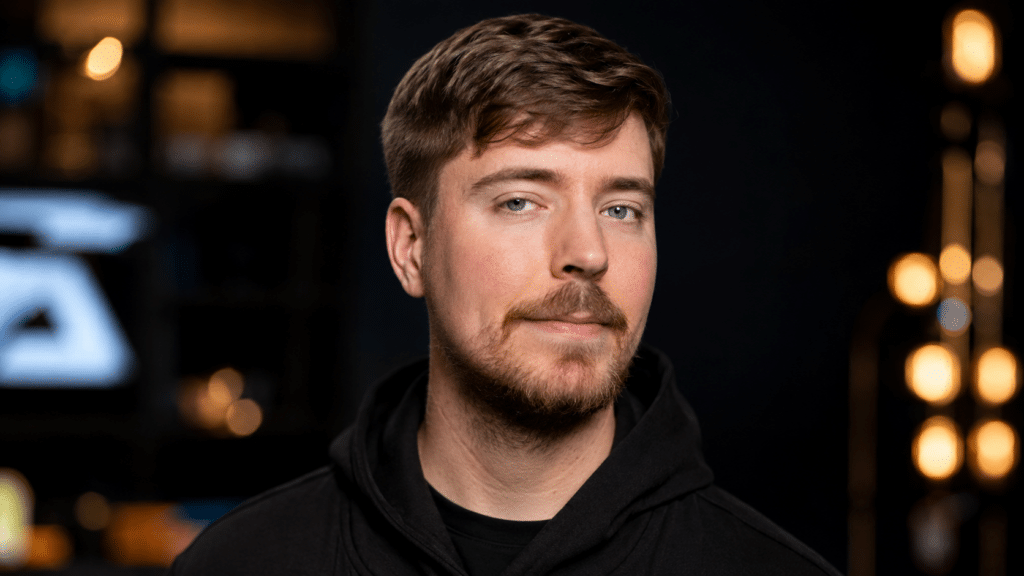 Close-up portrait of a man with short brown hair and a beard, wearing a black hoodie, against a dark studio backdrop with warm bokeh lights in the background.