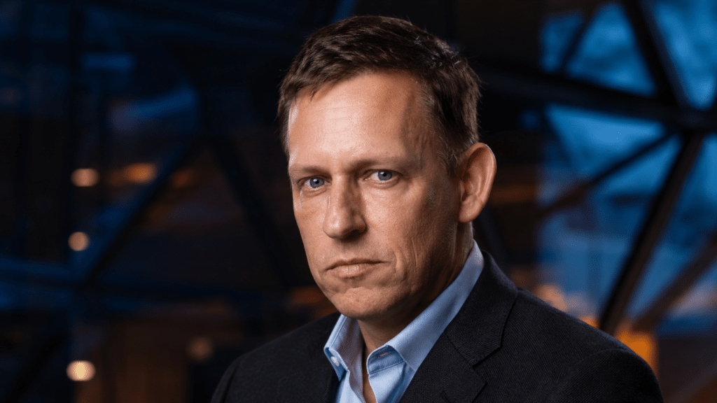 Professional headshot of a man in a dark blazer and light blue shirt, with a serious expression against a dark geometric background.
