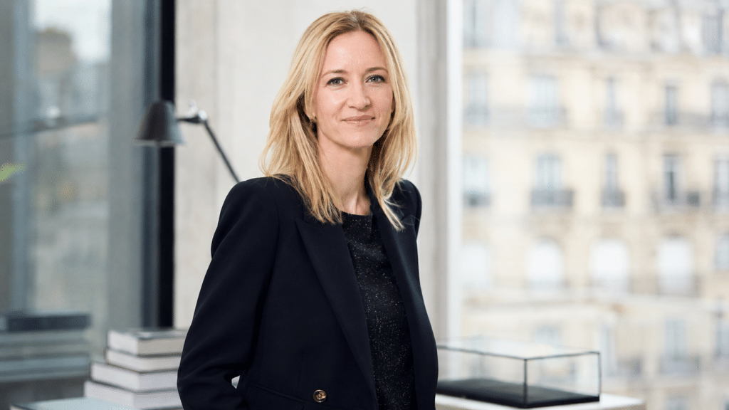 Professional woman with blonde hair in a black blazer, standing in a modern office with a cityscape view.